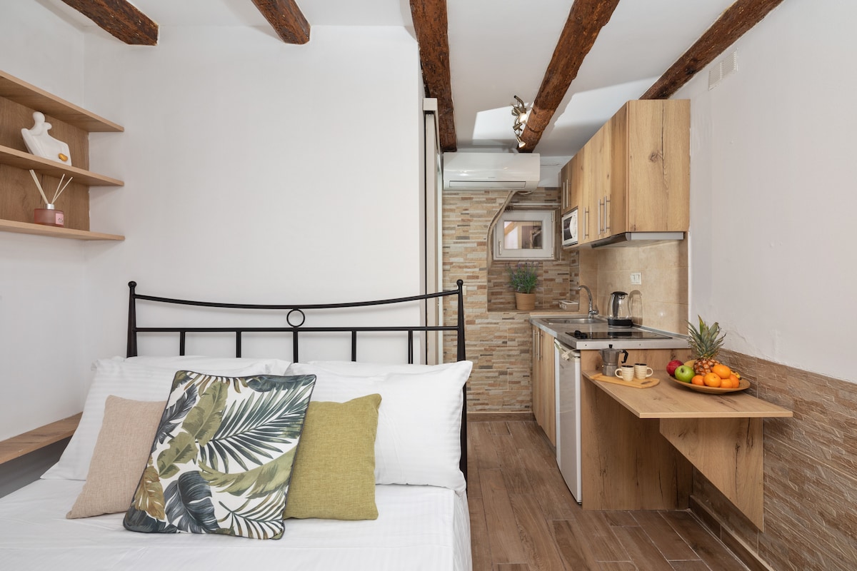 A bright and modern living space features a bed with soft white linens, accompanied by decorative pillows. In the background, a compact kitchen is visible with wooden cabinetry and a small dining table adorned with fresh fruits, emphasizing convenience and comfort.