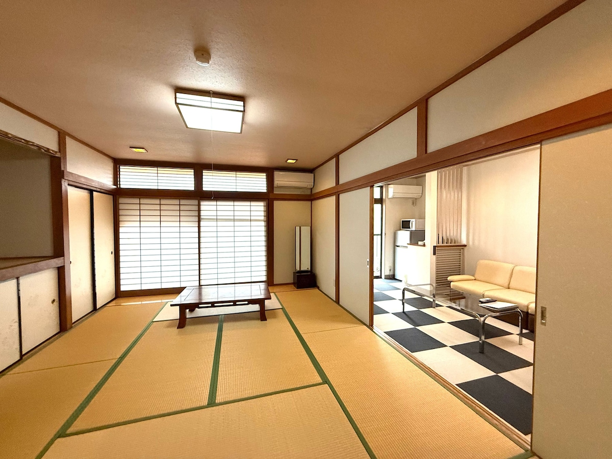 A spacious room features traditional Japanese tatami flooring and shoji screens allowing natural light to filter through. The area includes a low wooden table surrounded by seating. A doorway leads to a second room with modern seating and kitchenette amenities, highlighting the harmonious blend of styles.