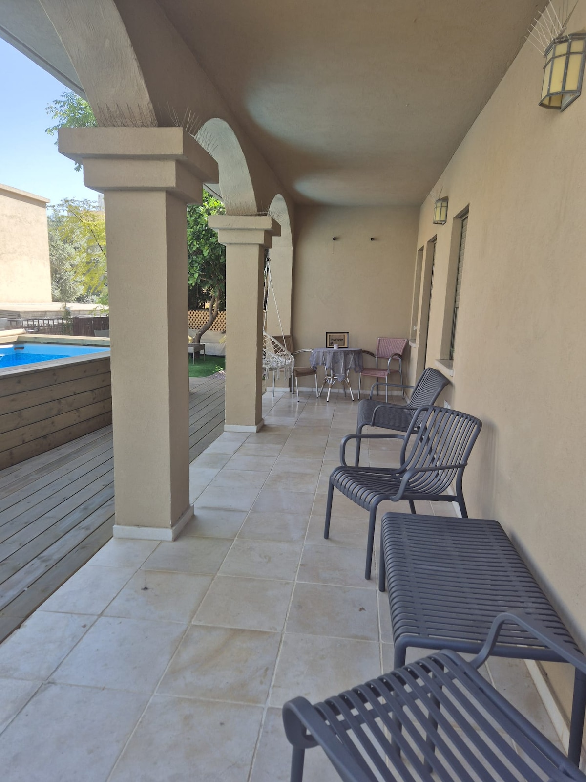 A covered patio features several metal chairs arranged along a tiled surface. The space is framed by arched columns and leads to a wooden deck and a private pool visible in the background, surrounded by greenery.
