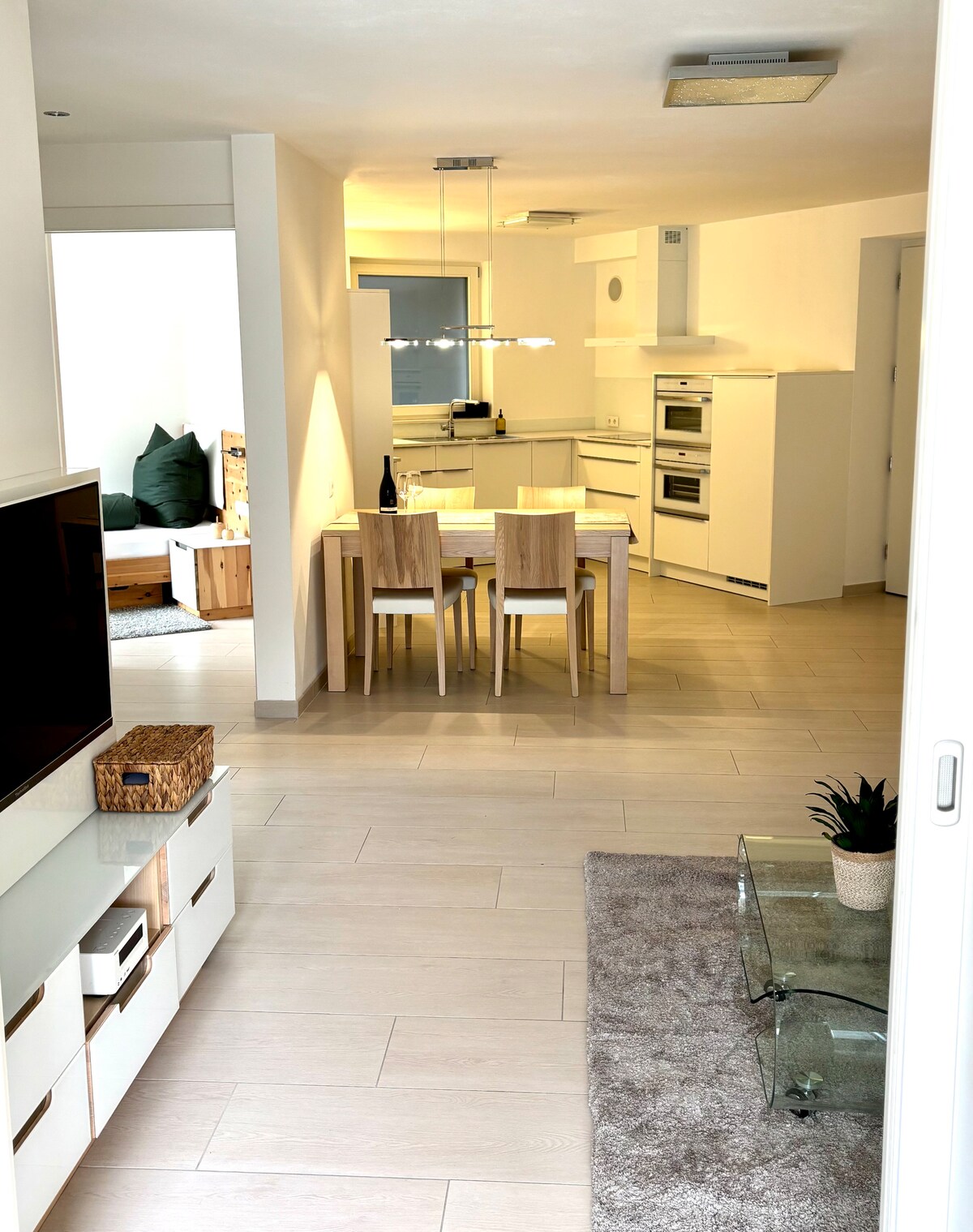 A bright and modern kitchen area is visible, featuring a dining table with four wooden chairs. The kitchen includes sleek cabinets and built-in appliances. Warm lighting enhances the inviting atmosphere, while a cozy lounge space is partially seen in the background.