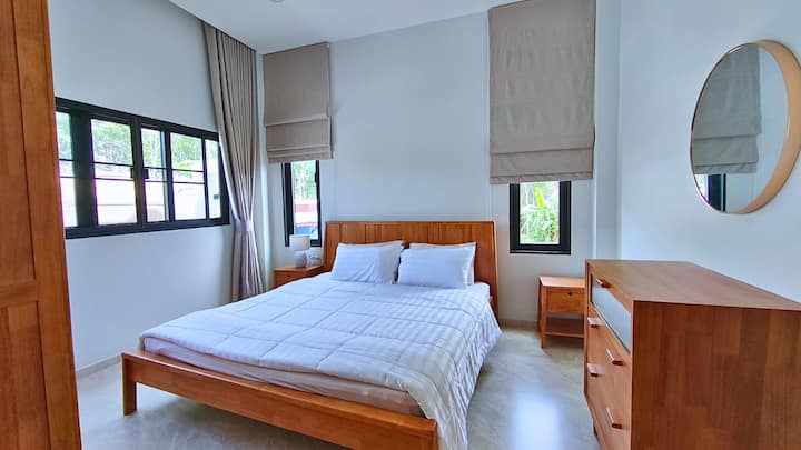 Bright and cozy bedroom with a comfortable double bed, fresh linens, and large wooden wardrobe. Minimalist design with natural light from two windows, creating a calm and relaxing atmosphere – perfect for restful nights.