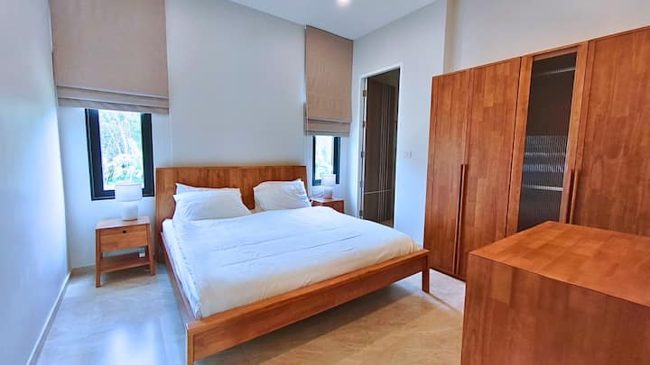 Bright and cozy bedroom with a comfortable double bed, fresh linens, and large wooden wardrobe. Minimalist design with natural light from two windows, creating a calm and relaxing atmosphere – perfect for restful nights.