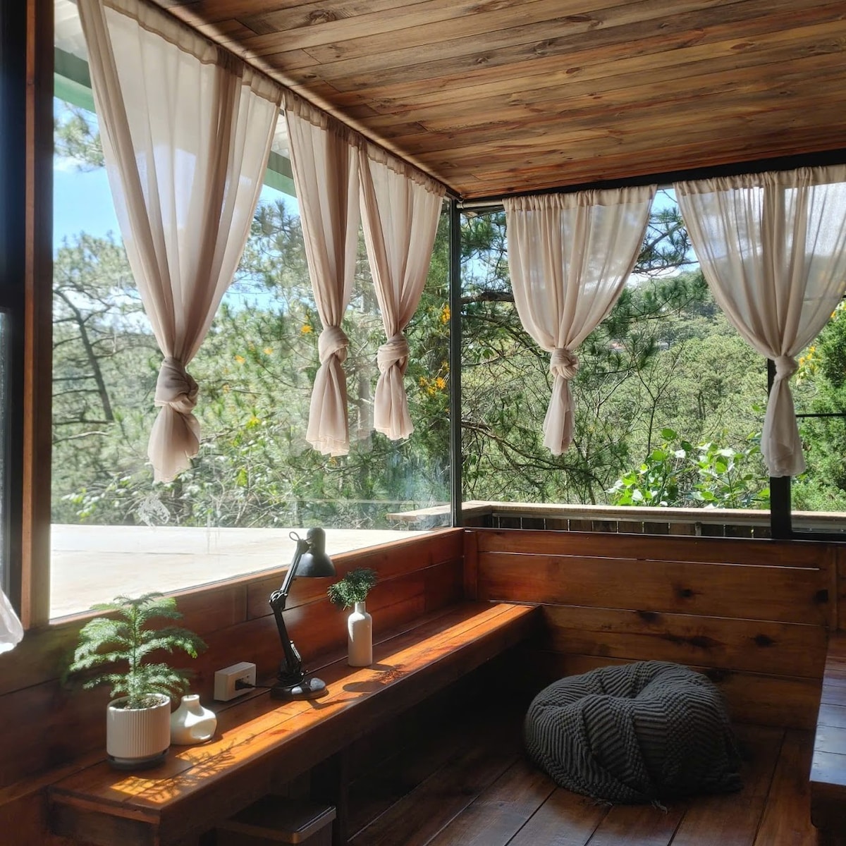 Natural light fills a cozy corner featuring soft, flowing curtains against wooden walls. A comfortable seating area is enhanced by a bench and a floor cushion. A small plant and a lamp sit on a side table, adding to the inviting simplicity of the space.