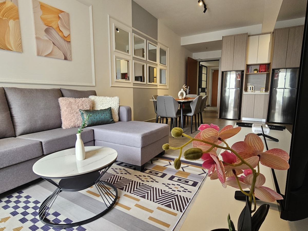 A cozy living space features a sectional sofa adorned with decorative cushions, complemented by a modern coffee table. A patterned area rug lies beneath, while a dining area with gray chairs and two refrigerator units can be seen in the background.