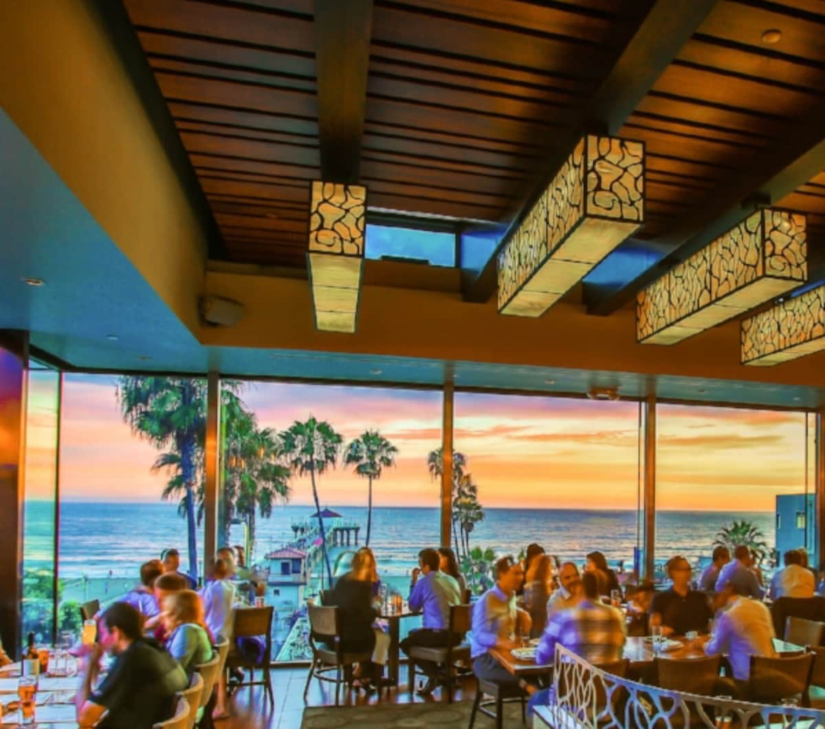 An inviting dining space is illuminated by unique pendant lights and features large windows that frame a vibrant sunset over the ocean. Guests are seen enjoying their meals while palm trees are visible outside, adding to the coastal ambiance.