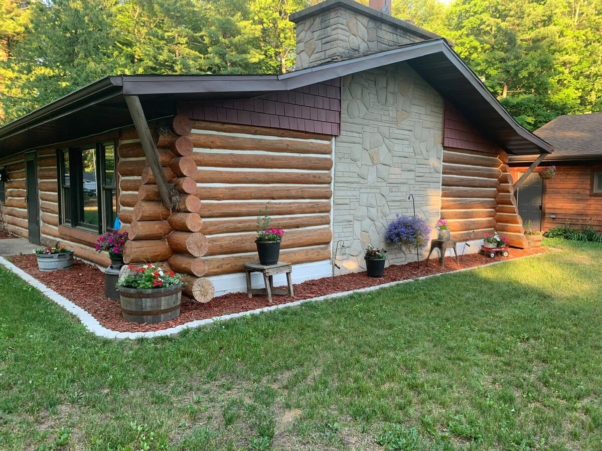 The exterior of the log cabin features a mix of natural wooden logs and stonework. Flower pots are placed around the entry, adding color to the landscape. A well-kept lawn surrounds the cabin, contributing to its homey environment.