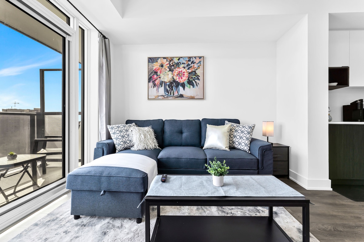 A modern living room features a dark blue sectional sofa with decorative pillows, positioned near a large window that allows natural light to fill the space. A dark coffee table sits in front of the sofa, complemented by a small potted plant, enhancing the clean, contemporary look.
