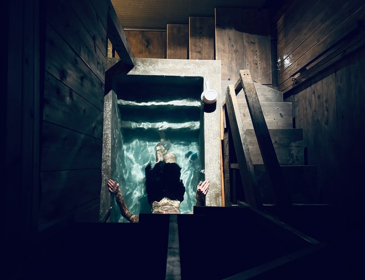 An indoor Finnish sauna is depicted, featuring a tranquil hot tub filled with clear water. Wooden walls and steps create a natural aesthetic. Soft overhead lighting contributes to a serene atmosphere, while an individual is seen enjoying the tranquility of the space.