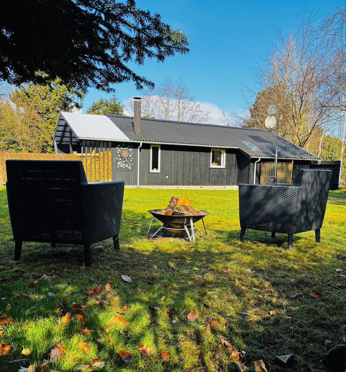 Two black wicker chairs are positioned around a circular fire pit in a grassy area. In the background, a dark wood cabin is situated under a clear blue sky, surrounded by trees. The setting offers a tranquil outdoor space for relaxation.