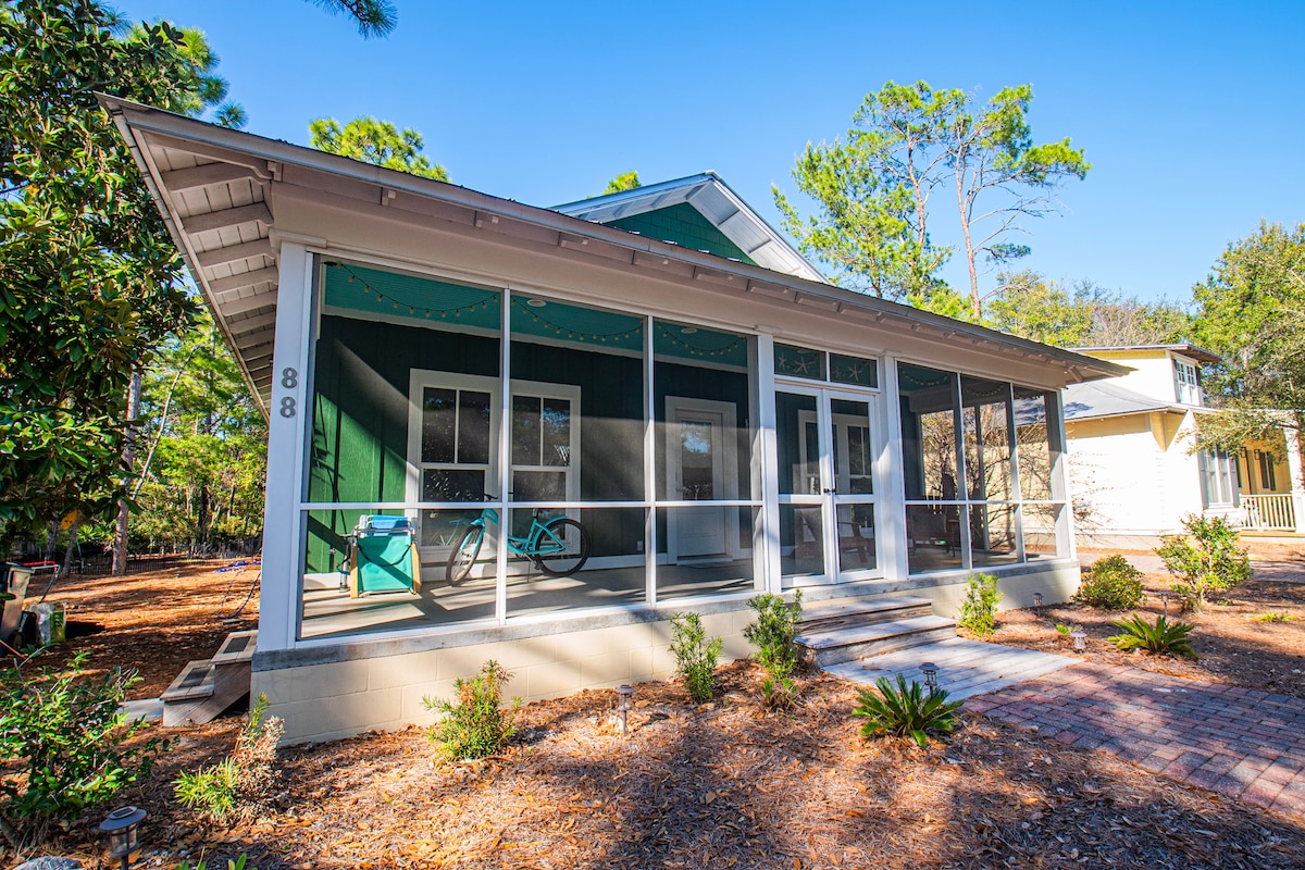 A charming single-family home is framed by a screened front porch, featuring multiple entry points and outdoor seating. Surrounding greenery and small shrubs add to the natural setting. The exterior is a blend of light and dark green hues, embraced by a well-maintained landscape.