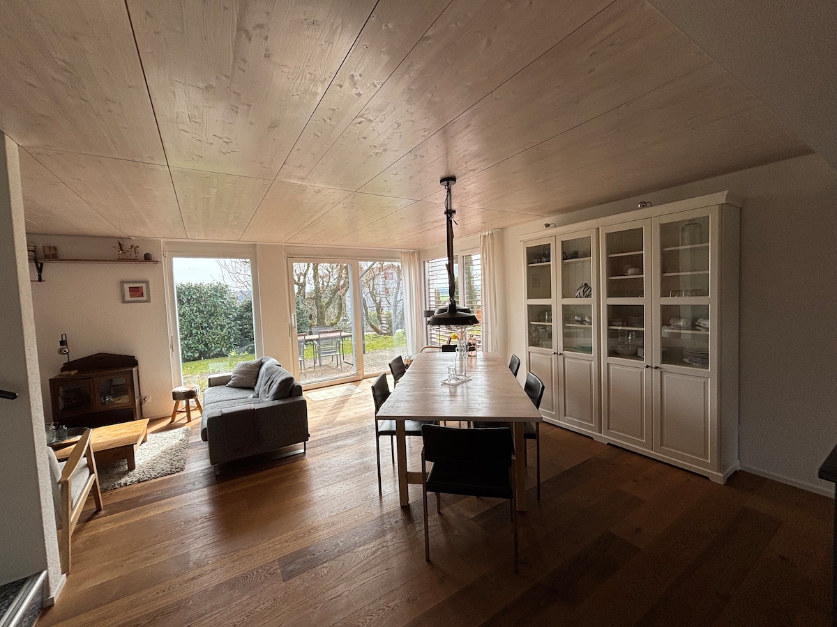A bright, open living space features large glass doors allowing natural light to flood in, revealing views of the garden. A stylish dining table is centrally positioned, surrounded by black chairs, while a cozy seating area with a grey couch and a coffee table invites relaxation.