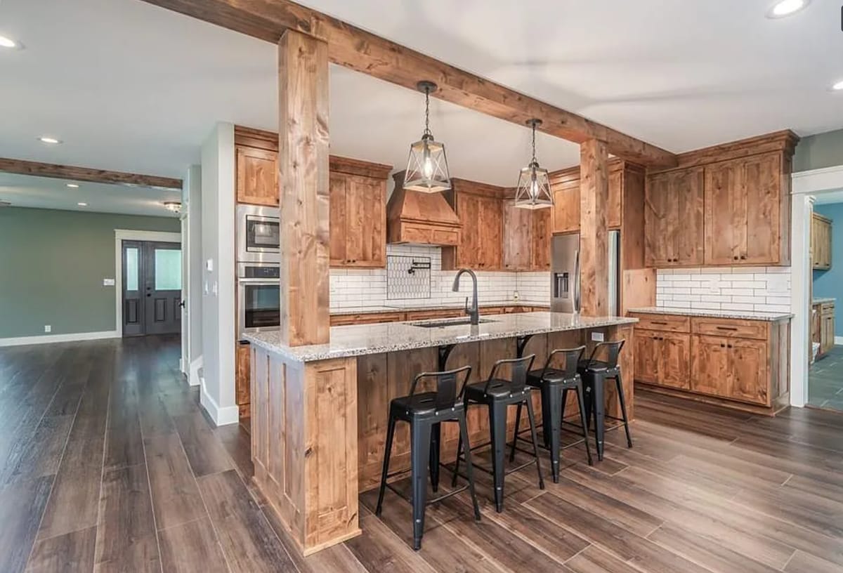 A modern kitchen is highlighted with warm wood cabinetry and a spacious island fitted with black metal stools. Natural light illuminates the area, showcasing the sleek countertop and the open layout leading to the adjoining living space.