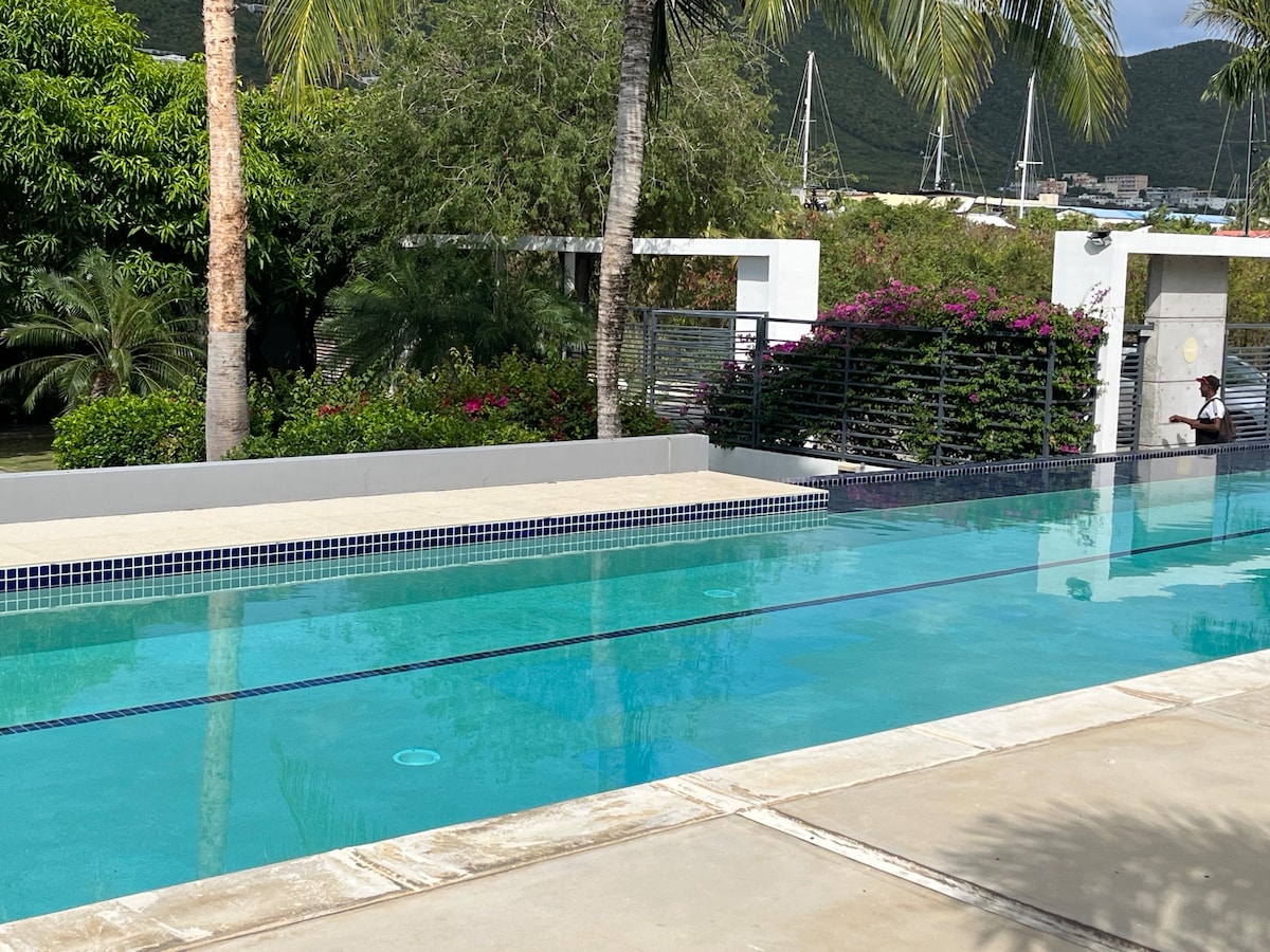 A tranquil infinity pool extends along the edge of a landscaped area, featuring lush greenery and flowering plants. The smooth water reflects the surrounding scenery, with palm trees framing the space. A serene atmosphere is created in this outdoor setting, ideal for relaxation.