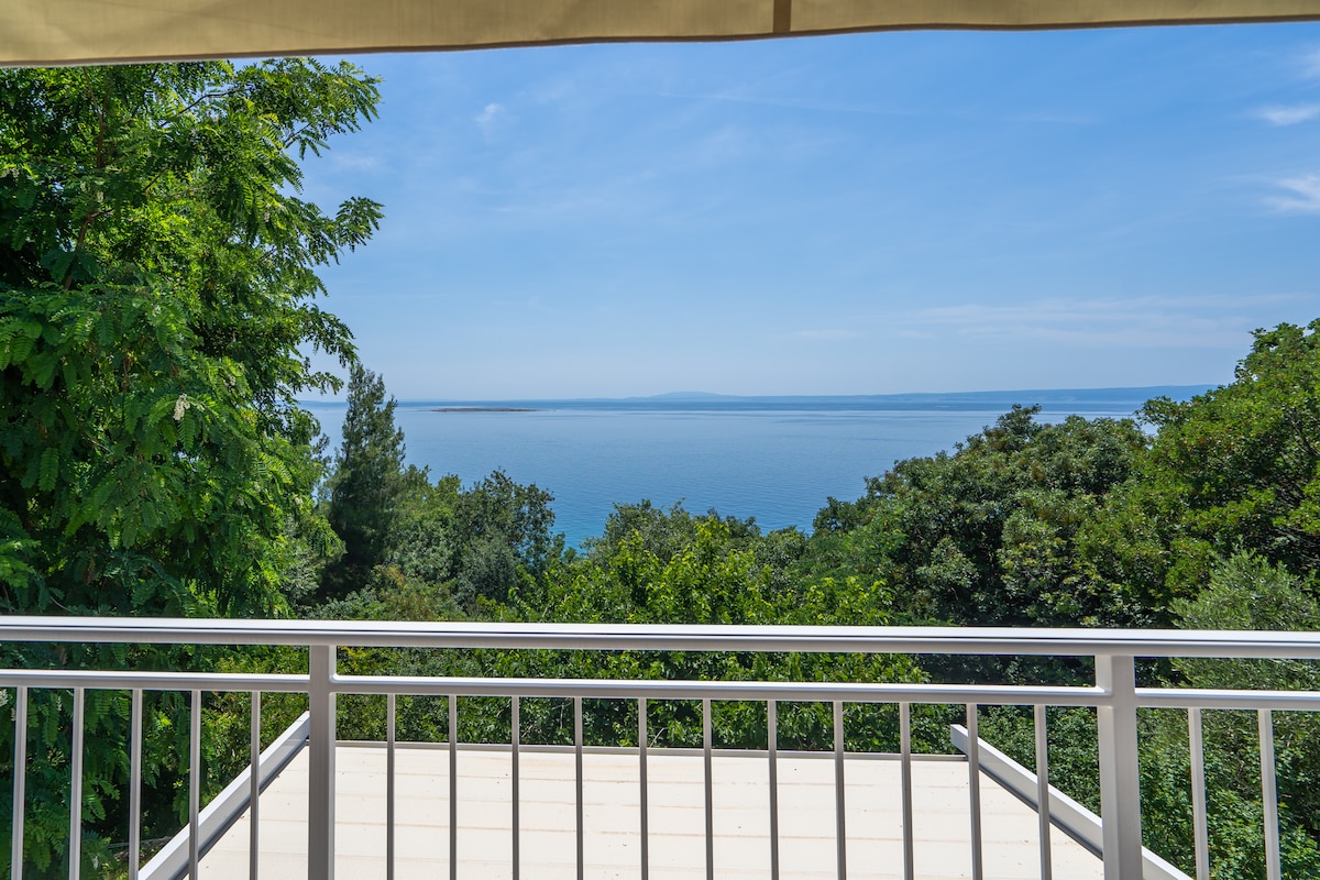 A clear view of the sea is presented, framed by lush greenery. The horizon meets the water in a gentle gradient of blues, under a bright sky with scattered clouds. A white railing offers a barrier to the outdoor space, enhancing the sense of an open vista.