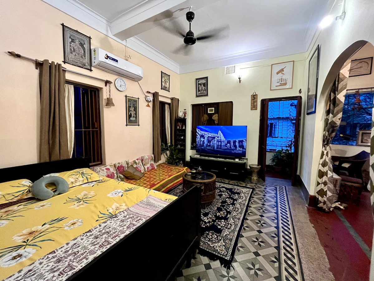 A spacious room features a mix of sleeping arrangements, including a double bed and a single bed, adorned with colorful bed linens. A large television stands opposite the beds, while the decor reflects traditional Bengali elements. Natural light filters in through curtained windows.