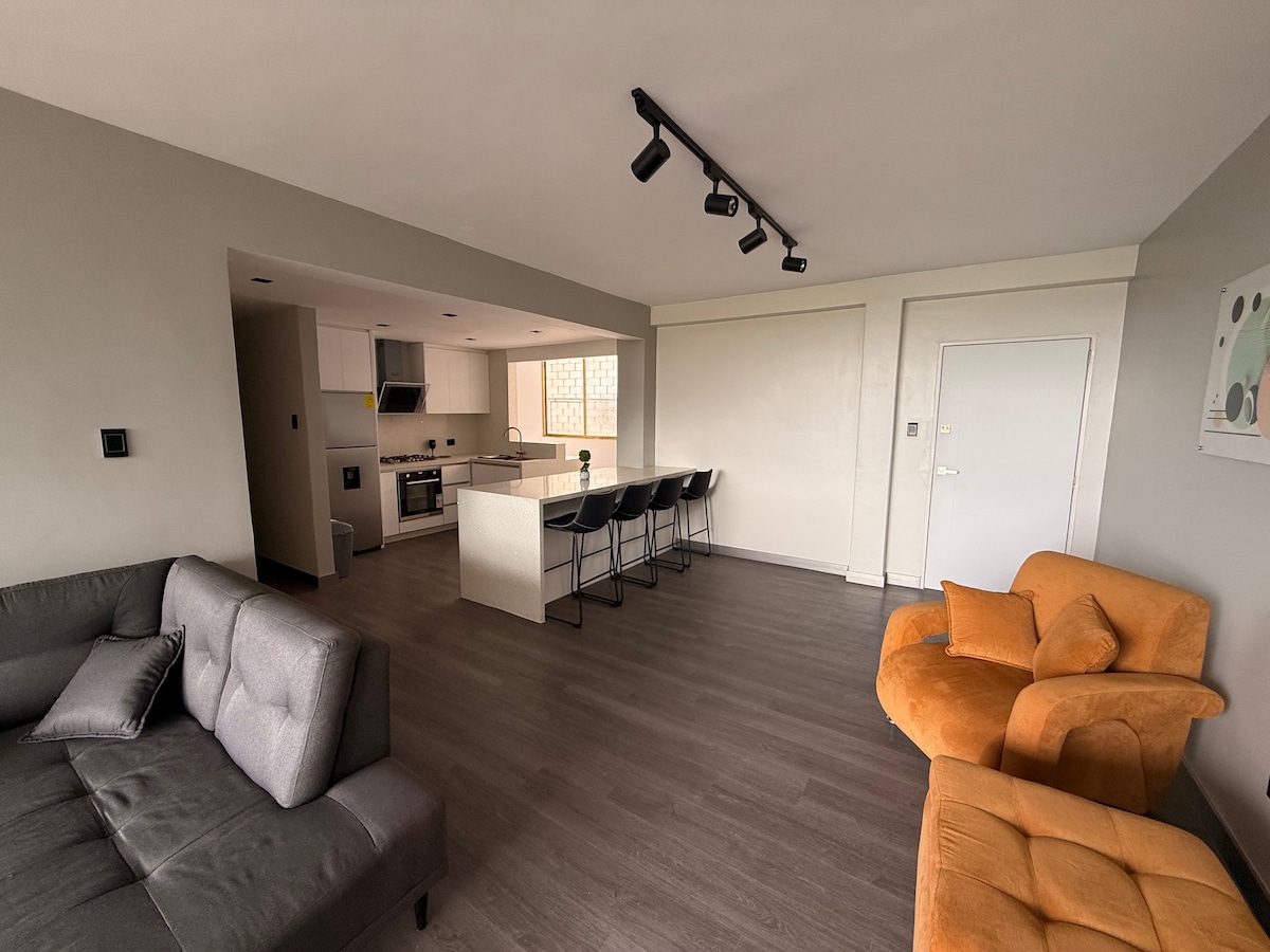An open-concept living space features a grey sofa and an orange armchair. A dining area with four barstools is positioned beside a modern kitchen, equipped with essential appliances. Natural light enters through a large window, illuminating the sleek flooring and neutral walls.