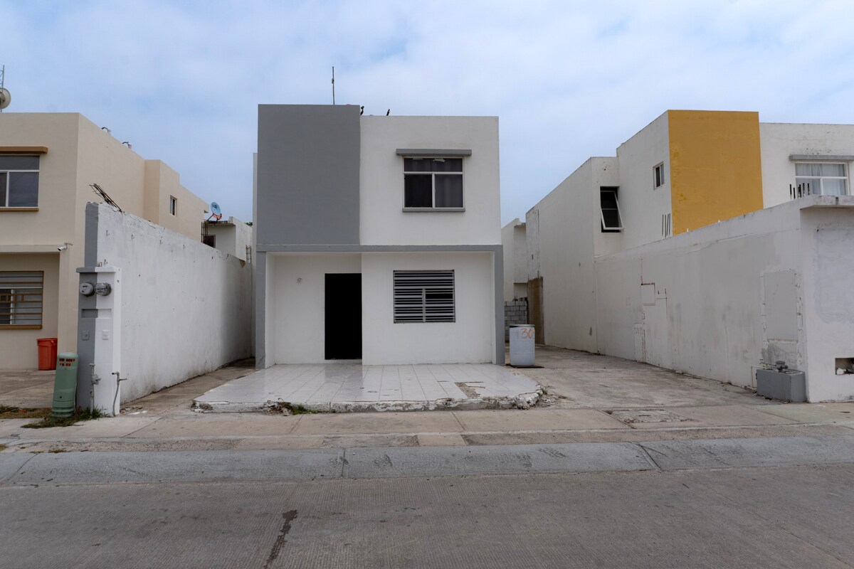 A modern two-story house is depicted with a minimalist design, featuring a gray and white exterior. Large windows provide natural light to the interior. The surrounding area includes nearby structures and a paved street, with an open space in front of the house.