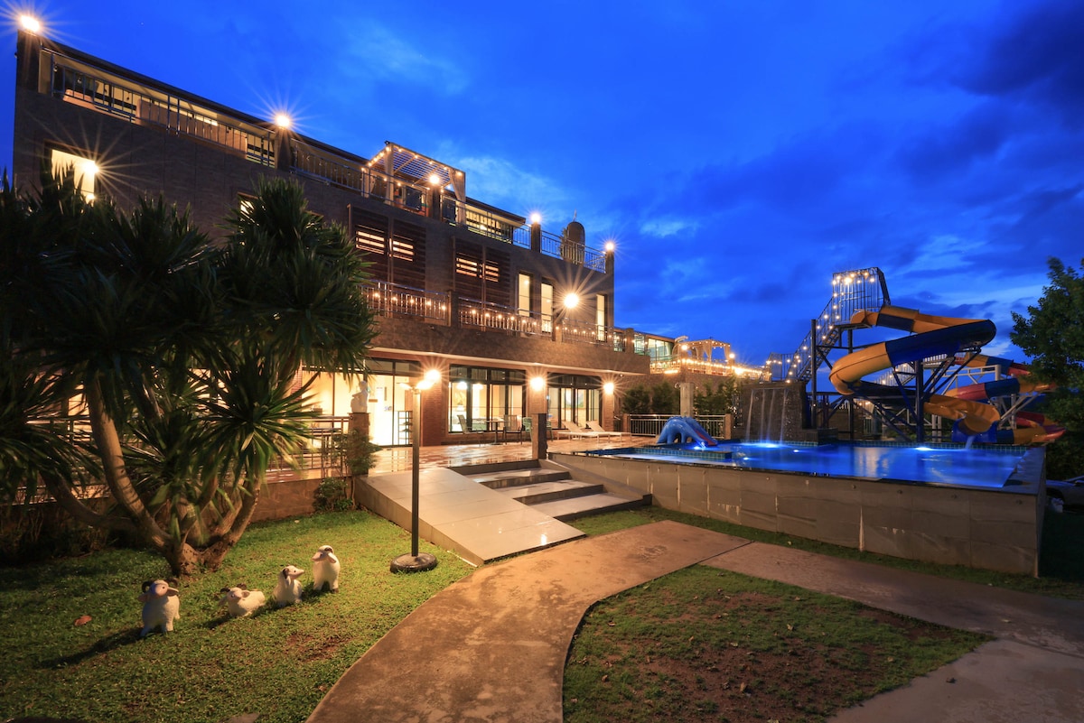 A well-lit outdoor area showcases a modern building with multiple levels. A pool and water slide are visible, along with a manicured lawn featuring decorative plants. Pathways meander through the space, highlighting various seating areas and soft lighting illuminating the scene.