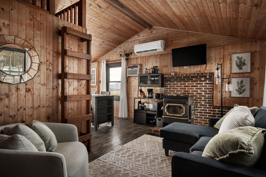 The cozy living area features wood-paneled walls and a comfortable seating arrangement, including a sectional sofa and armchair. A flat-screen TV is mounted on the wall above a brick fireplace. Shelving holds decorative items and a mini bar setup is visible in the background.