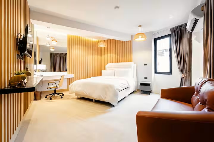 A private room features a spacious bed with crisp white linens against a backdrop of warm striped walls. A comfortable armchair sits adjacent to a window that provides natural light, while a small desk and a flat-screen TV complete the cozy atmosphere.