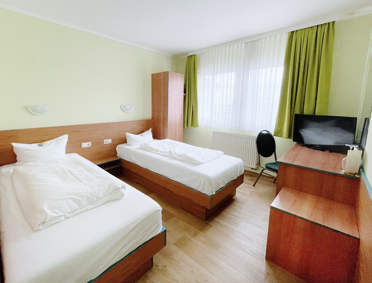 A well-lit guest room features two comfortable single beds with white bedding, a wooden dresser, and a sleek desk. A chair is positioned by the desk, while a television is mounted on the wall. Natural light enters through large windows adorned with light green curtains.