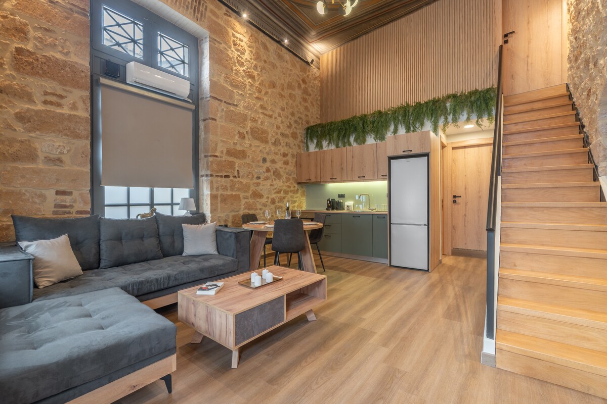 A spacious living area features stone walls and wooden accents, complemented by hardwood flooring. A comfortable sectional sofa is paired with a wooden coffee table. An open kitchen area is visible with modern appliances, while a staircase leads to the upper level.