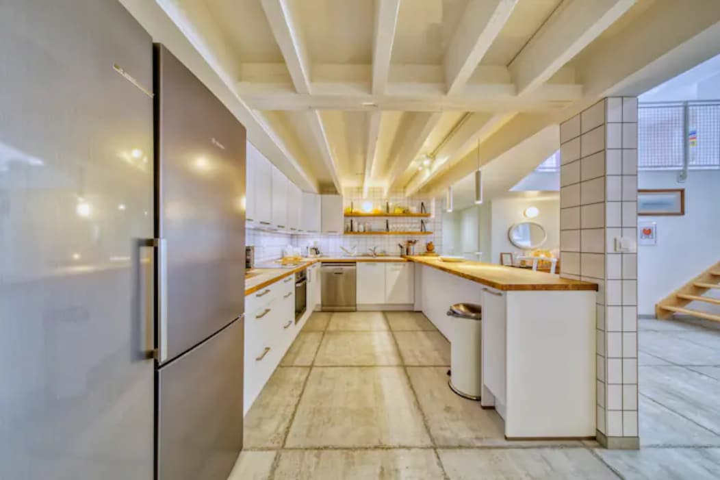 A modern kitchen is equipped with sleek cabinets and ample counter space. Stainless steel appliances are integrated, including a refrigerator and oven. Exposed wooden beams are visible above, and natural light brightens the open area. A waste bin is positioned near the cabinetry.