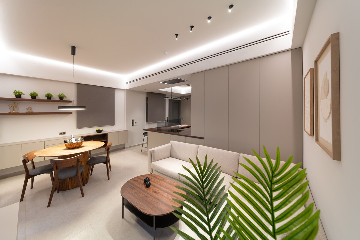 A modern living area is shown, featuring a light-colored sofa and a round wooden dining table surrounded by chairs. The space is enhanced by subtle greenery and sleek cabinetry, with soft lighting highlighting the open kitchen in the background.