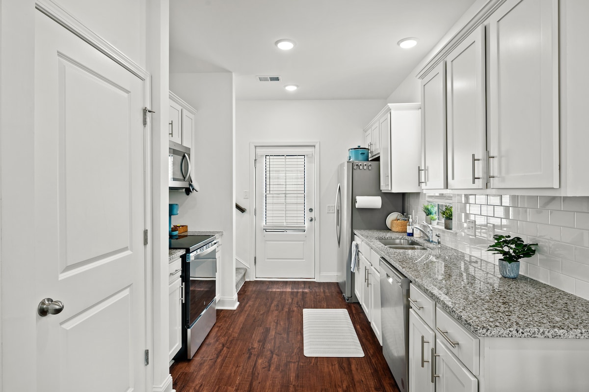 A modern kitchen features sleek white cabinetry and stainless steel appliances. The granite countertops provide ample workspace, complemented by a double sink. A door leads outside to a small patio area, while natural light illuminates the space through a nearby window.