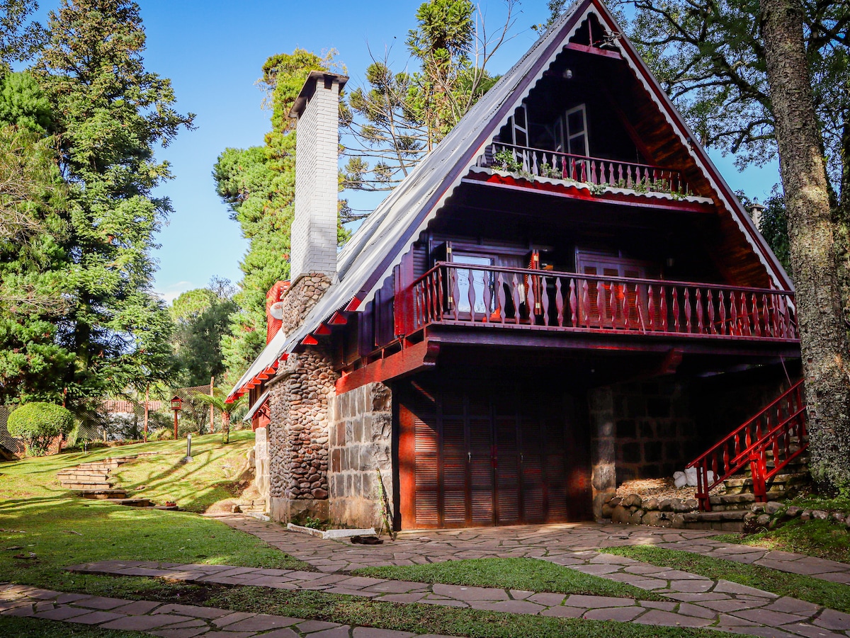 The exterior of the cabin is highlighted by its rustic stone walls and red wooden accents. A sloped roof adds character, while a charming balcony overlooks the manicured garden. Tall trees and greenery surround the cabin, enhancing its serene and nature-oriented environment.