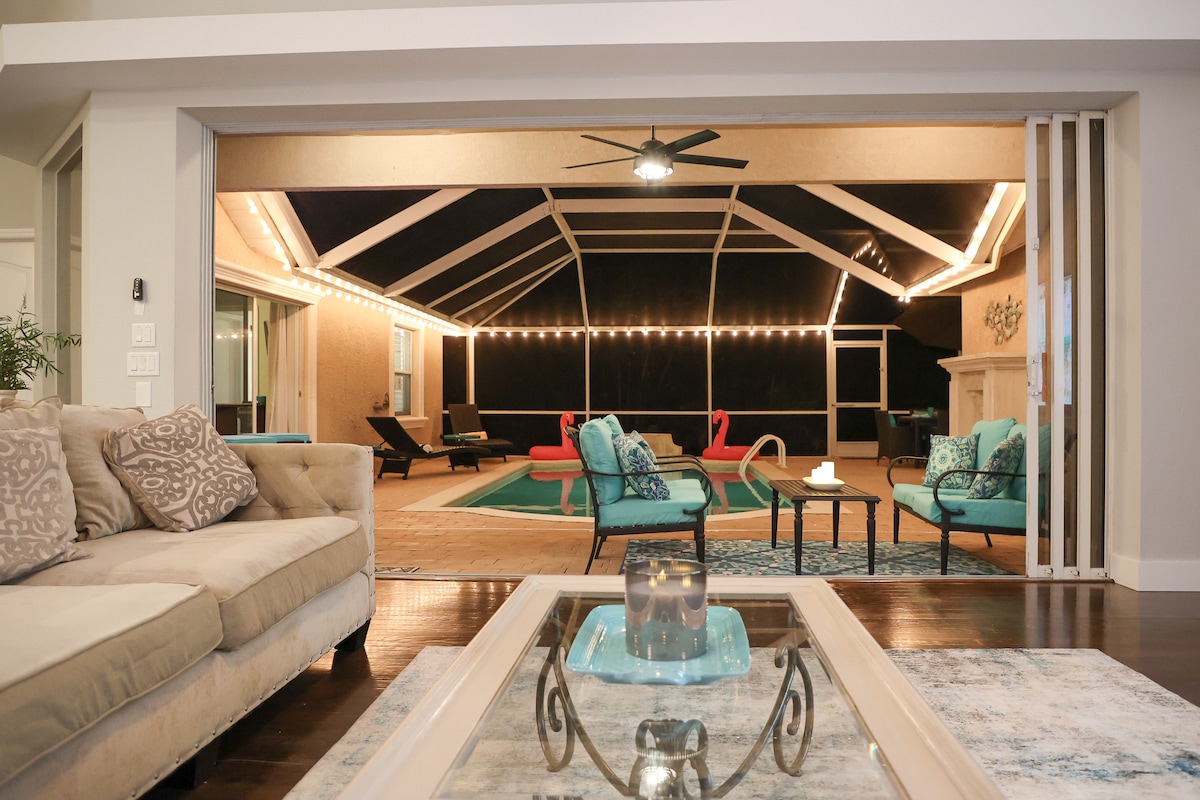 A spacious living area features a comfortable sectional sofa, a glass coffee table, and colorful decorative pillows. Large glass doors open to the screened lanai, revealing a heated pool with lounge chairs and ambient lighting, blending indoor and outdoor relaxation spaces.