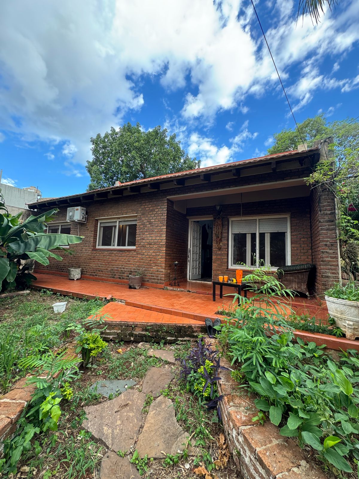 A spacious brick house is surrounded by lush greenery, featuring a wooden deck that connects to a well-maintained garden. The exterior includes a large window, allowing natural light to enter. A small table is set on the porch, inviting relaxation in a serene environment.
