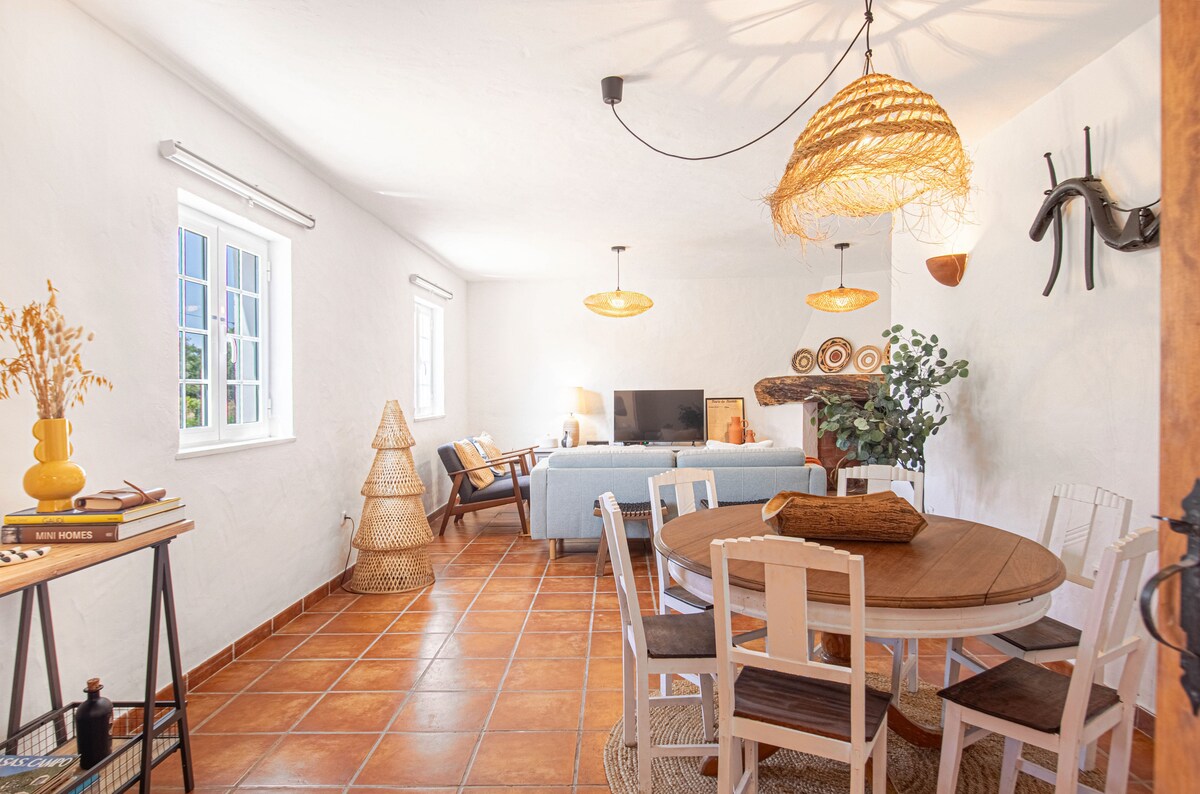 A bright living area features a combination of natural light and neutral tones, accented by woven light fixtures. A cozy blue sofa is positioned near a television, while a round wooden dining table is surrounded by white chairs. Decorative elements include potted greenery and rustic wall art.