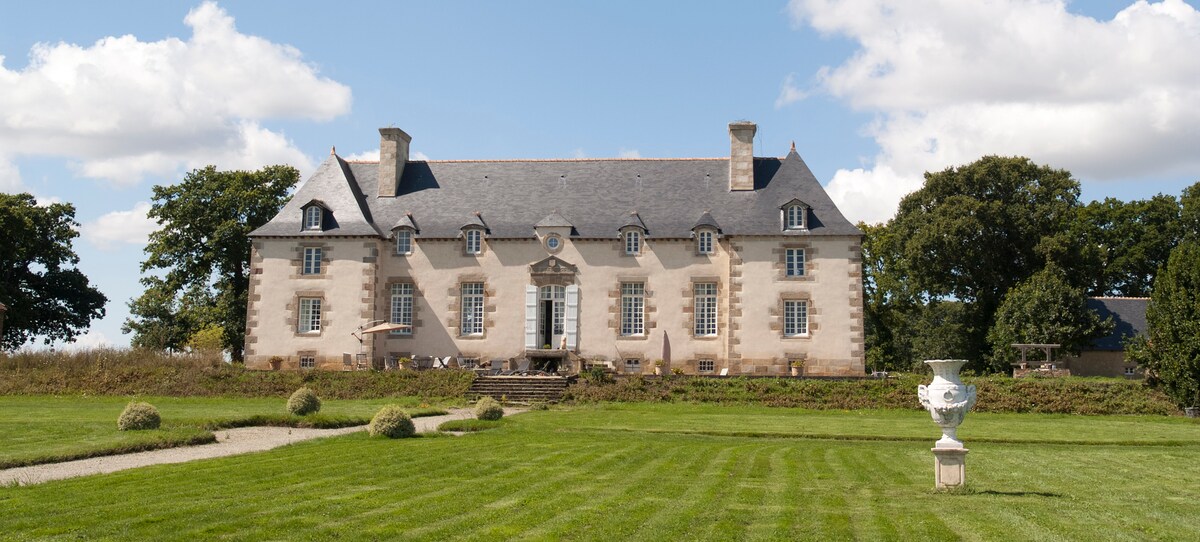 The exterior of a restored manor house is displayed amid a lush green landscape. The building features multiple gabled roofs, large windows, and a central entrance with an ornamental door. A well-maintained lawn leads up to the structure, framed by trees and bushes.