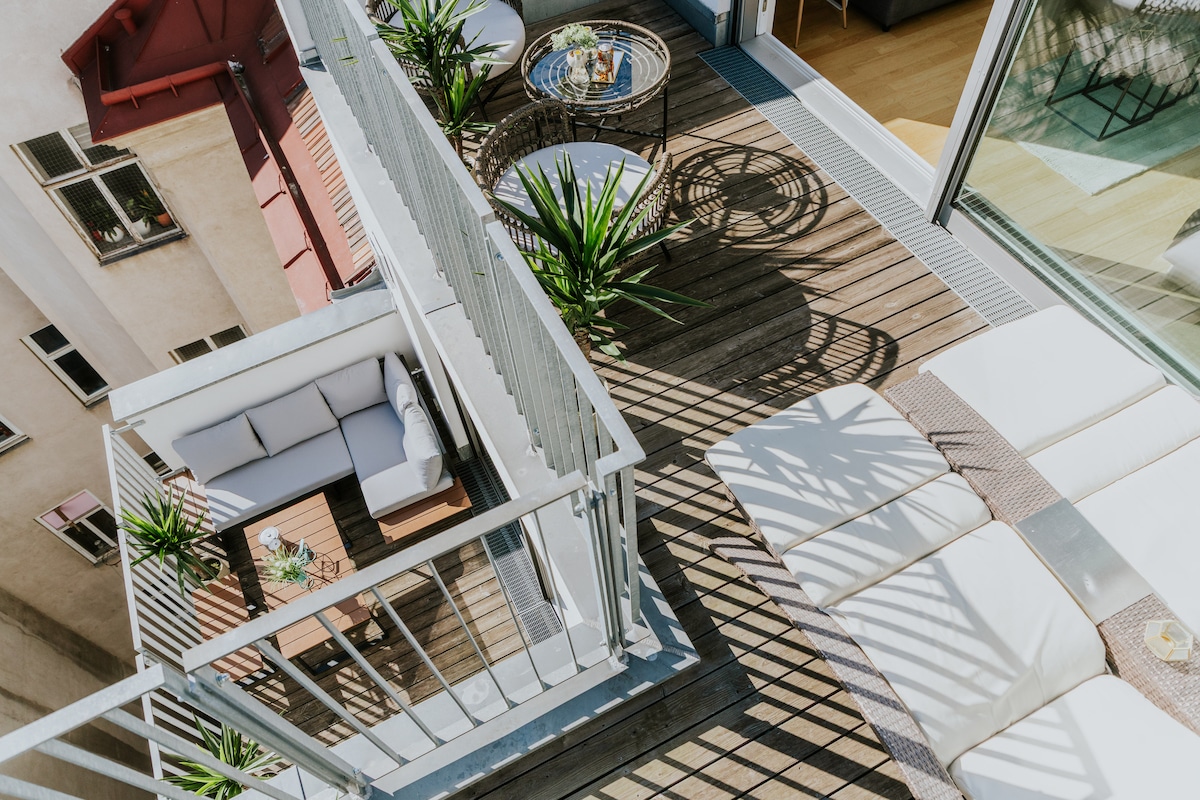 A spacious balcony features a wooden deck with several potted plants. Comfortable seating is arranged around a circular table, providing a cozy outdoor area. Sunlight casts intriguing shadows across the space, enhancing the inviting layout for relaxation.
