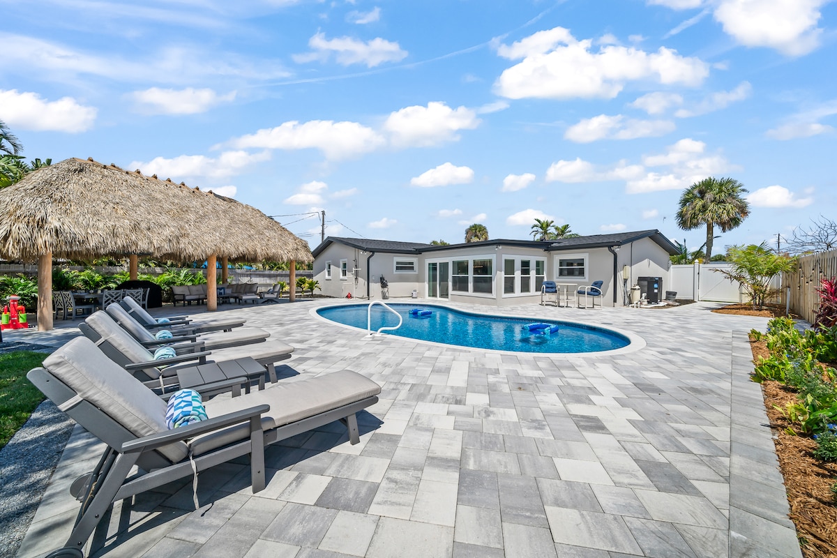 A spacious outdoor area features a saltwater pool surrounded by a patio, enhanced by sun loungers and a large tiki hut. The tiki hut offers shaded outdoor dining and lounging space, with tropical landscaping complemented by clear blue skies.