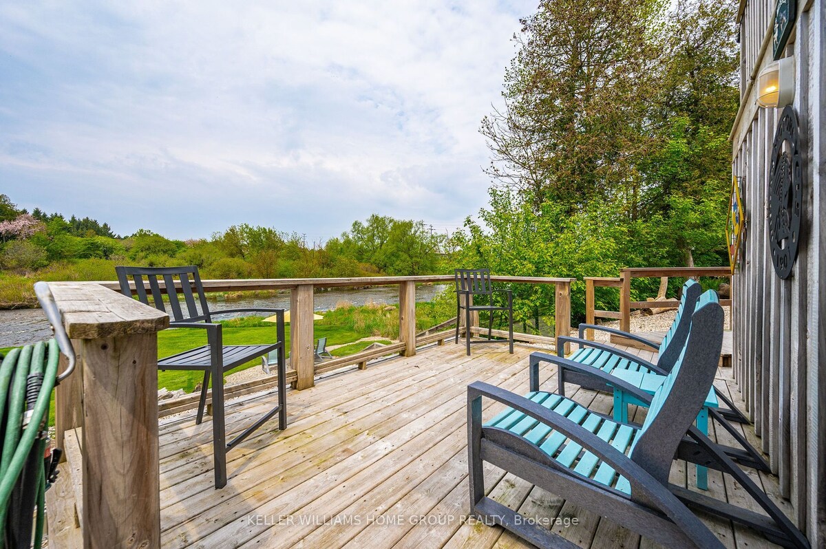 A wooden deck overlooks the Grand River, featuring several seating options including two Adirondack chairs and barstools. Lush greenery surrounds the area, contributing to a peaceful outdoor setting. The space invites relaxation with ample room for guests to enjoy the tranquil views.