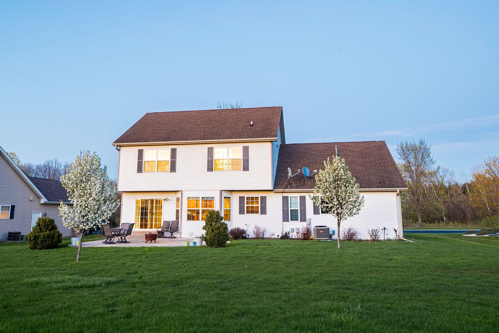 A two-story house is depicted from the backyard, featuring a well-maintained lawn. The exterior is clad in white siding, with large windows reflecting the evening light. Trees with blossoms frame the patio area, enhancing the outdoor space's inviting atmosphere.