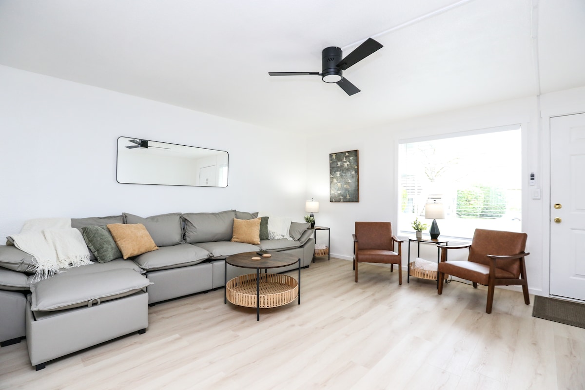 A contemporary living area features a sectional sofa with neutral upholstery and decorative pillows, accompanied by a round coffee table. Two leather chairs provide additional seating, while natural light enters through large windows, illuminating the space. A mirror hangs on the wall, enhancing the room's openness.