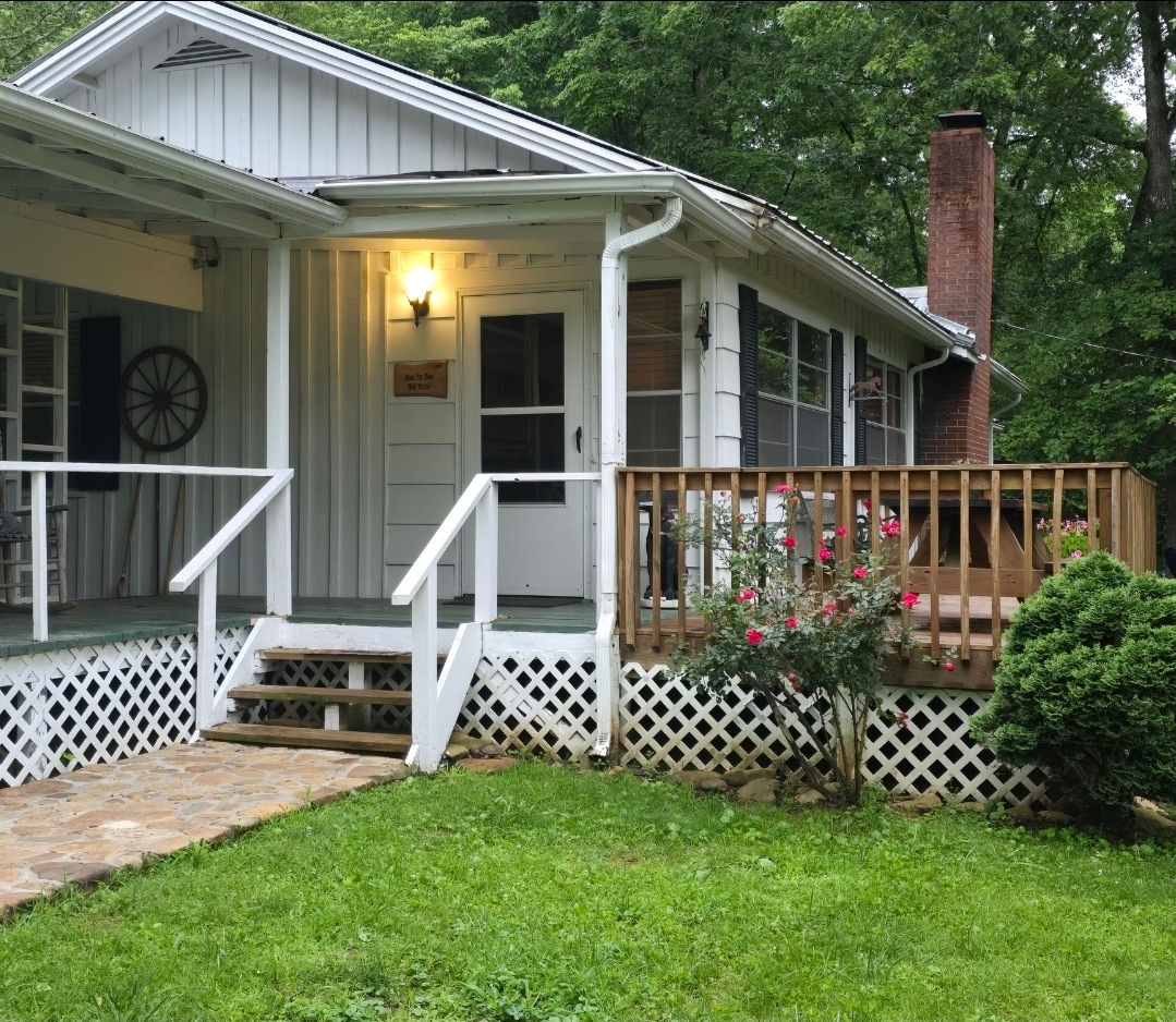 The exterior of a charming family home is presented, featuring a welcoming front porch with wooden steps leading to the entrance. A small wooden deck is visible, adorned with a couple of blooming rose bushes and bordered by greenery, creating a serene setting.
