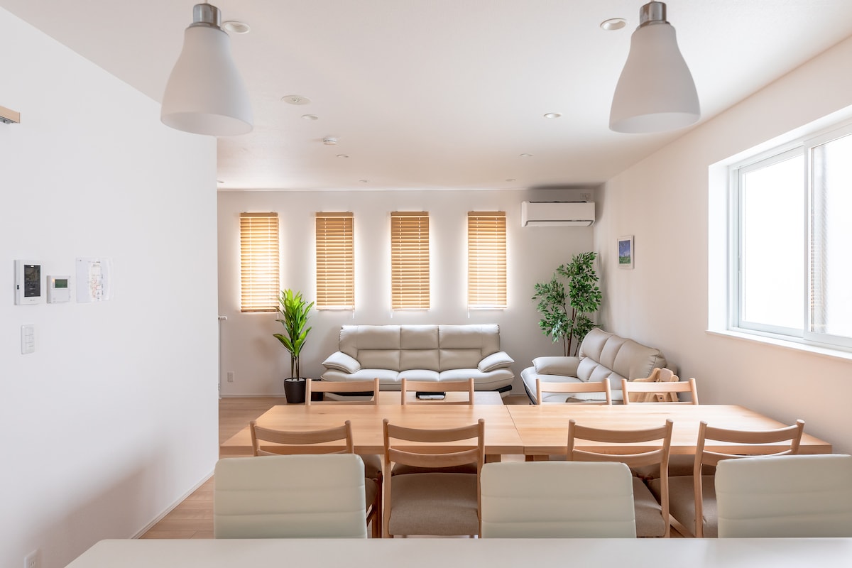 A bright and airy living space is highlighted by several windows with wooden blinds that provide natural light. Comfortable seating arrangements are visible, including a sectional sofa and armchairs. A dining table with multiple chairs occupies the foreground, complemented by a potted plant in one corner.
