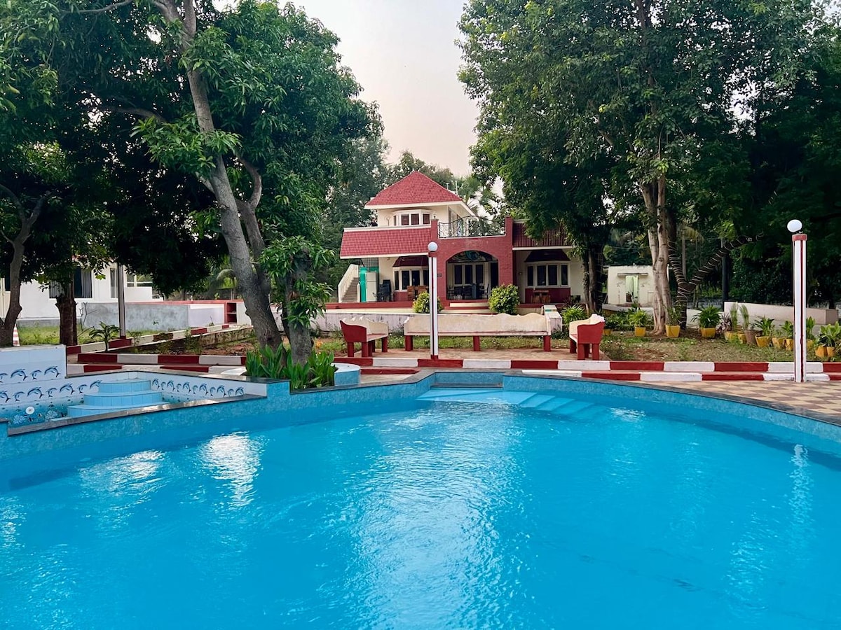 A clear blue swimming pool reflects the surrounding greenery, with wooden benches positioned along its edge. In the background, a charming red-roofed building is partially obscured by trees, offering a serene and inviting atmosphere to the setting.