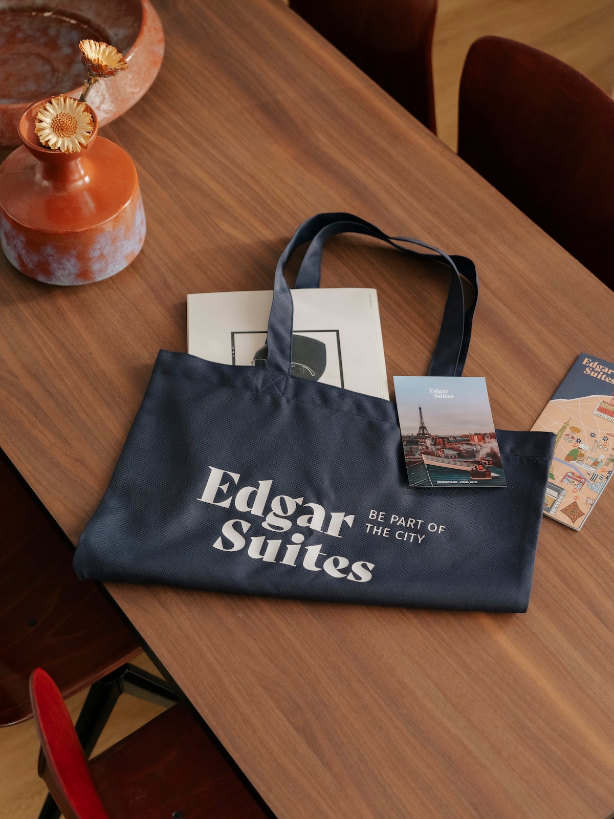 A dark blue tote bag with the text 'Edgar Suites' and the phrase 'BE PART OF THE CITY' is placed on a wooden dining table. Nearby, a brochure and a small framed piece of art complement the scene, along with an ornamental vase featuring a floral design.