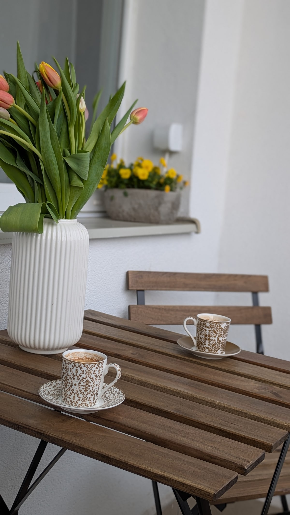 A small outdoor balcony table is set for two, featuring a pair of coffee cups on decorative saucers. A tall, white vase filled with fresh tulips and a pot of yellow flowers are positioned nearby, bringing a touch of nature to the space.