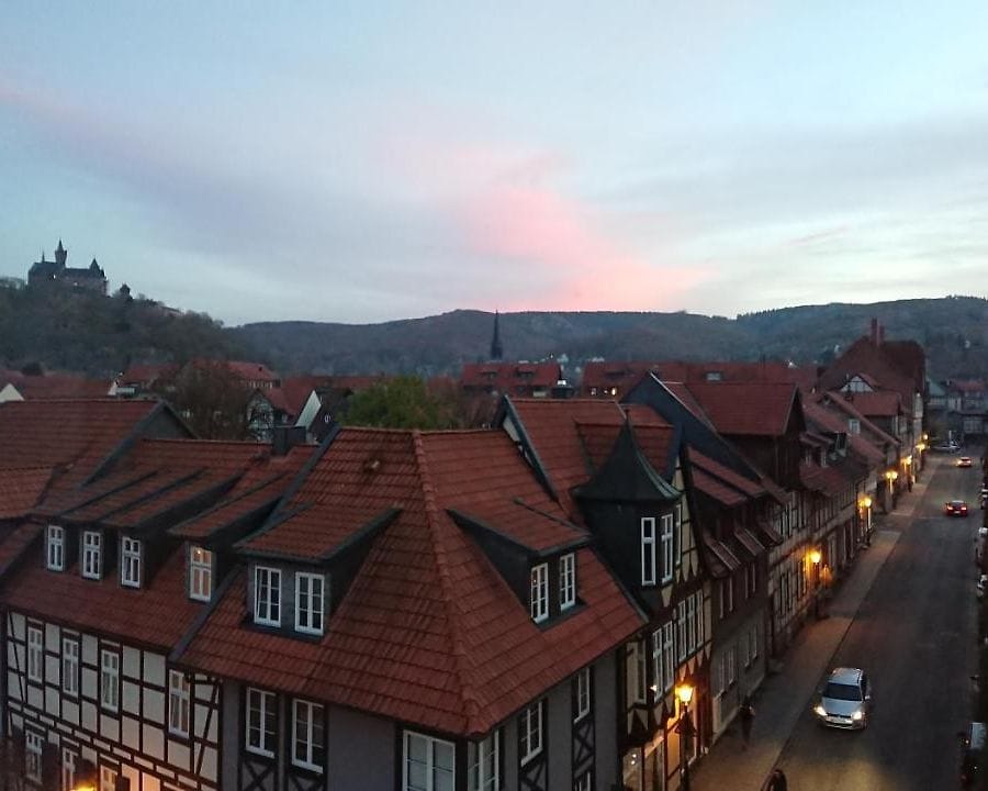 An overview of the historic town features charming timber-framed buildings with red roofs lining the street. A gentle evening sky transitions from soft orange to light blue, framing hills in the background. The scene reflects a peaceful atmosphere in a quaint setting.