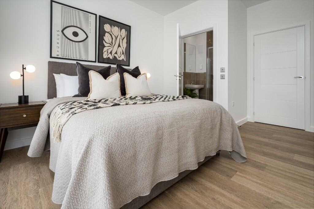 A spacious bedroom features a comfortable bed with soft linens and decorative pillows. A bedside table with a stylish lamp is positioned next to the bed. The room is brightened by natural light coming from the doorway leading to an attached bathroom.