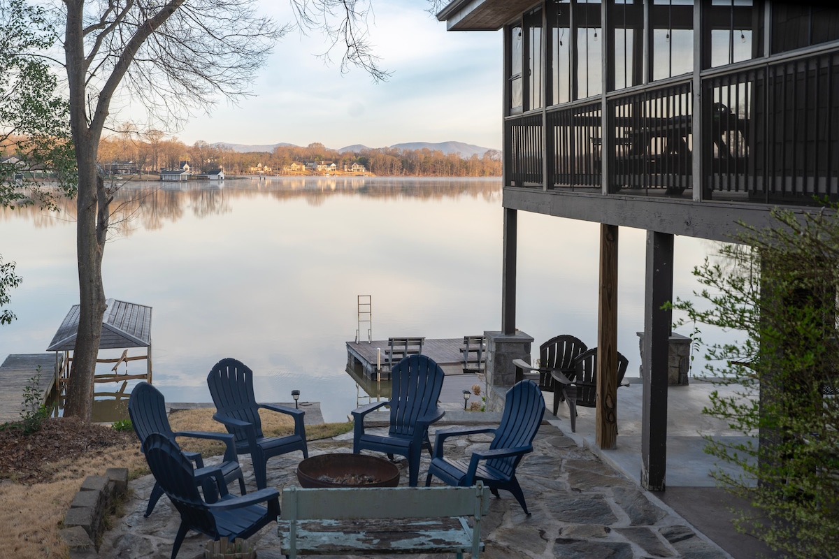 An outdoor fire pit area is surrounded by four blue Adirondack chairs on a stone patio, overlooking a serene lake. The calm water reflects the surrounding trees and distant houses, while a dock extends into the lake, inviting tranquility.
