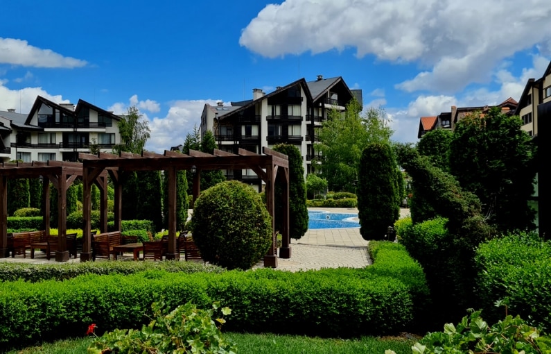 A well-maintained outdoor area features manicured hedges and a collection of trees. A wooden pergola stands with several benches arranged underneath. A clear swimming pool is visible in the background, surrounded by a tiled patio, while clouds drift across the blue sky.