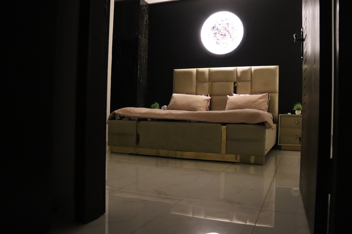 A dimly lit bedroom is seen from the doorway, featuring a king-sized bed with a neutral-toned coverlet. A unique moon design is showcased on the dark wall above the bed. Glossy tiles reflect light, enhancing the room's contemporary decor.