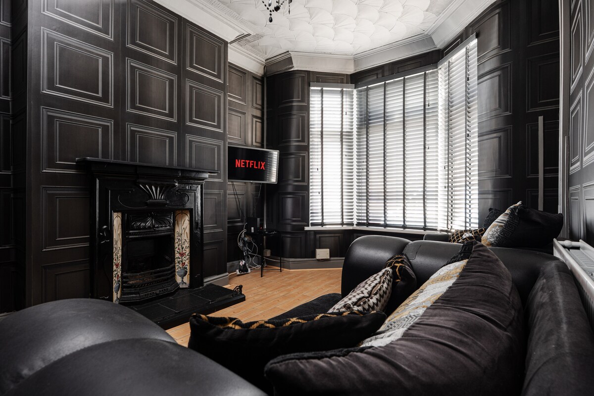 A spacious living area showcases bold black walls and a bay window, allowing natural light to fill the room. A modern television is positioned for viewing, complemented by a large sectional sofa adorned with patterned cushions. A decorative fireplace adds character to the space.